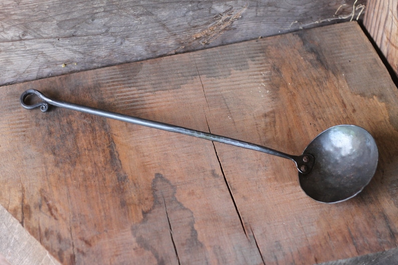 May include: A black metal ladle with a long handle and a rounded bowl. The handle has a decorative loop at the end.