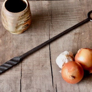 May include: A black metal fireplace poker with a twisted handle and a forked end. The poker is lying on a wooden surface with three onions and a head of garlic.