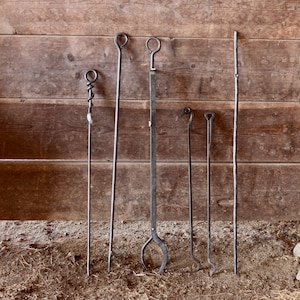 May include: A set of six fireplace tools with various handles, including a poker, a shovel, and a tongs. The tools are made of metal and have a rustic finish. They are hanging on a wooden wall.