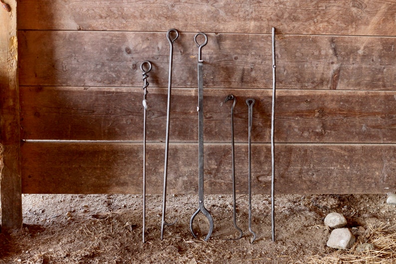 May include: A set of six fireplace tools with various handles, including a poker, a shovel, and a tongs. The tools are made of metal and have a rustic finish. They are hanging on a wooden wall.