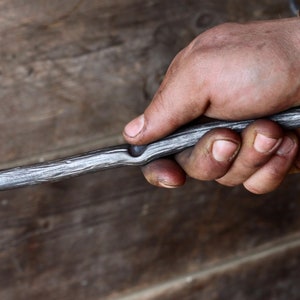 May include: A hand holds a long, thin, metal rod with a slight bend in the middle. The rod has a rough, textured surface and appears to be made of iron.