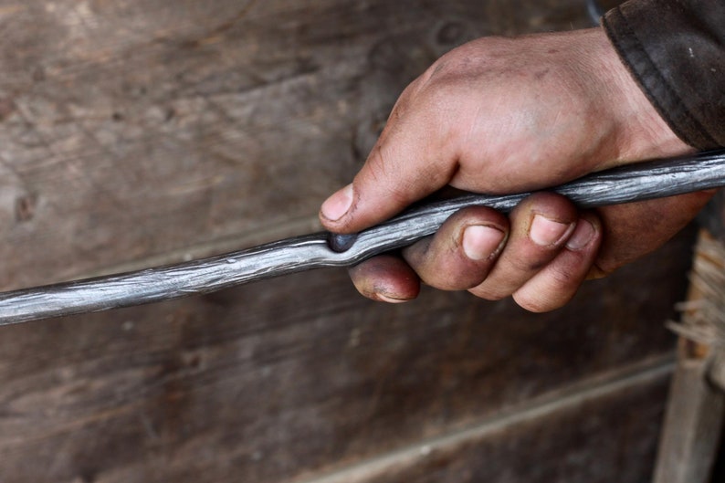 May include: A hand holds a long, thin, metal rod with a slight bend in the middle. The rod has a rough, textured surface and appears to be made of iron.