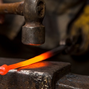 May include: A blacksmith uses a hammer to strike a glowing red metal rod on an anvil.