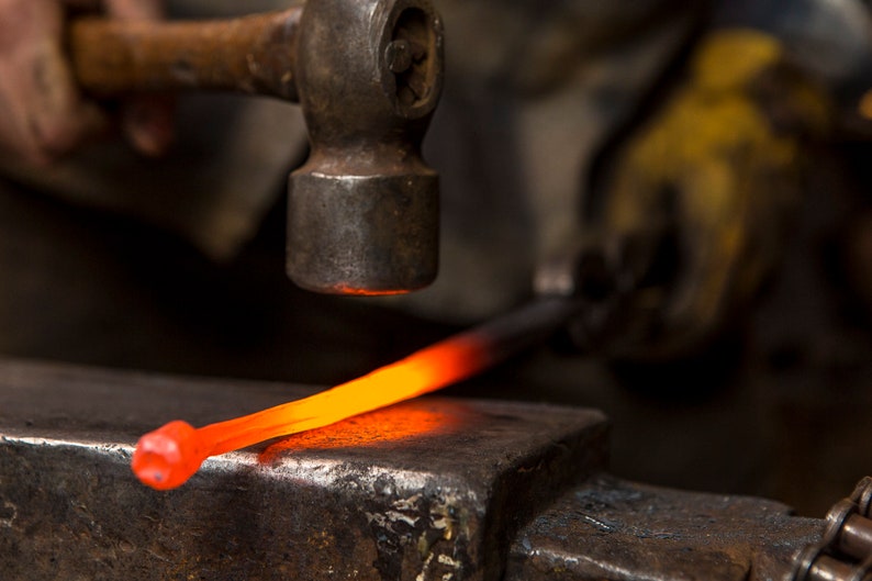 May include: A blacksmith uses a hammer to strike a glowing red metal rod on an anvil.