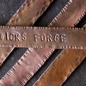 May include: Close-up of three hammered copper metal tags with a scalloped edge. The top tag has the words "Wicks Forge" stamped into the metal.