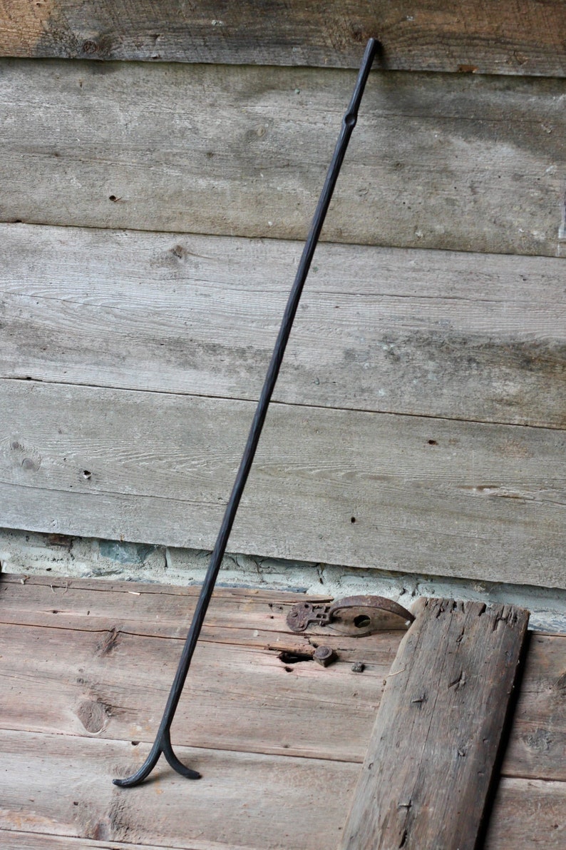May include: A long, black metal fireplace poker with a forked end. The poker is leaning against a wooden wall.