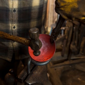 May include: A person using a hammer to shape a red metal bowl held in a vise. The bowl is being worked on by a person wearing a yellow leather glove.