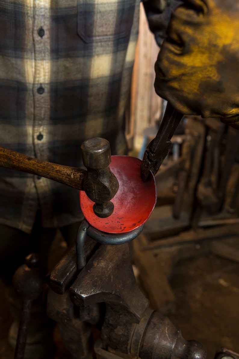 May include: A person using a hammer to shape a red metal bowl held in a vise. The bowl is being worked on by a person wearing a yellow leather glove.