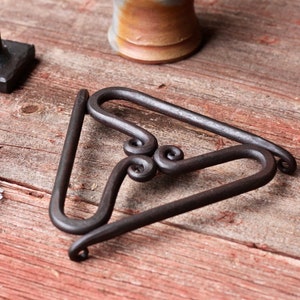 May include: A black metal trivet with a triangular shape and three decorative swirls. The trivet is made of wrought iron and has a rustic look.