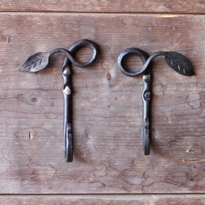 May include: Two black metal hooks with leaf details, mounted on a wooden surface.