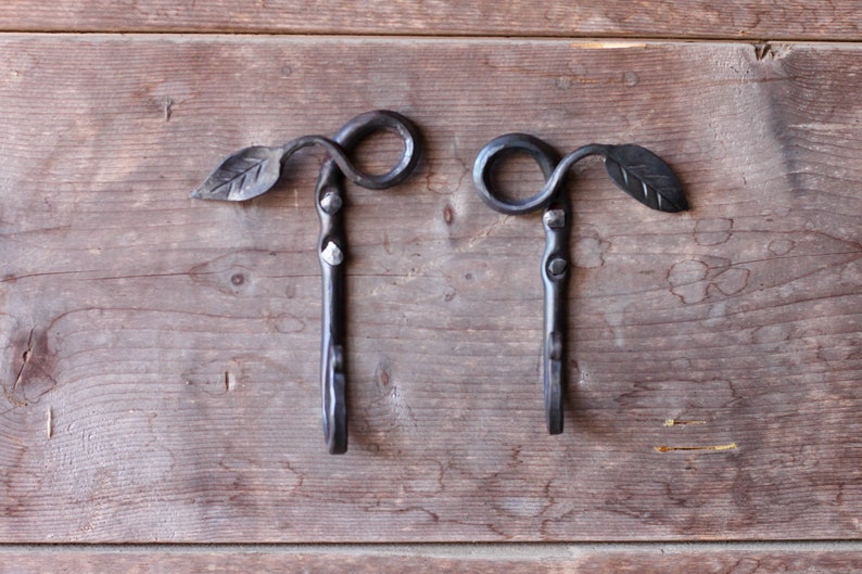 May include: Two black metal hooks with leaf details, mounted on a wooden surface.