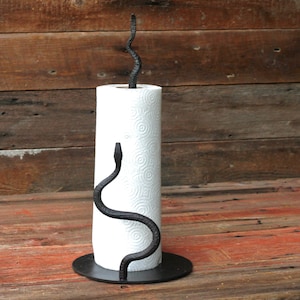 May include: Black metal snake-shaped paper towel holder with a white paper towel roll. The holder is on a wooden surface.