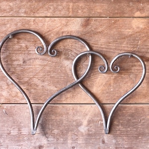 May include: Two interlocking heart-shaped sculptures crafted from dark gray metal. Each heart features a smooth, curved outline with decorative scrollwork at the top center, creating a continuous, flowing design that connects the two individual pieces.