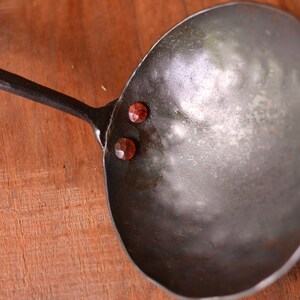 May include: A large, metal ladle with a long, hammered handle. The ladle has three red rivets on the side.