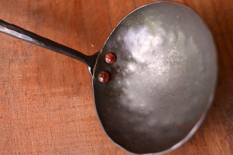 May include: A large, metal ladle with a long, hammered handle. The ladle has three red rivets on the side.