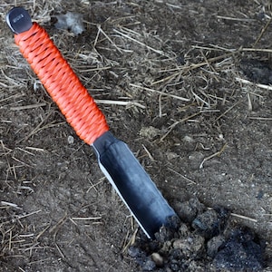 May include: A small, hand-held garden trowel with a black metal blade and a bright orange woven handle. The trowel is partially buried in dark soil, suggesting use in gardening or landscaping.