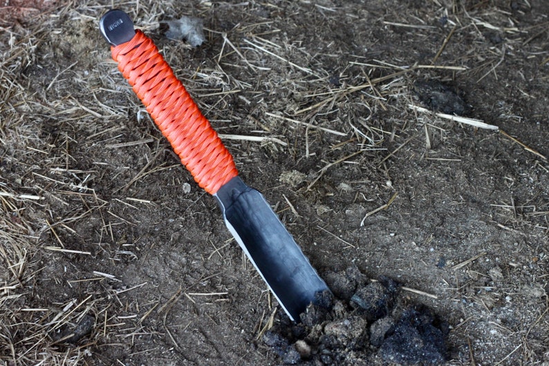 May include: A small, hand-held garden trowel with a black metal blade and a bright orange woven handle. The trowel is partially buried in dark soil, suggesting use in gardening or landscaping.