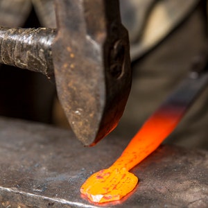 May include: A close-up of a blacksmith hammering a piece of red-hot metal on an anvil. The metal is glowing orange and is being shaped into a point.