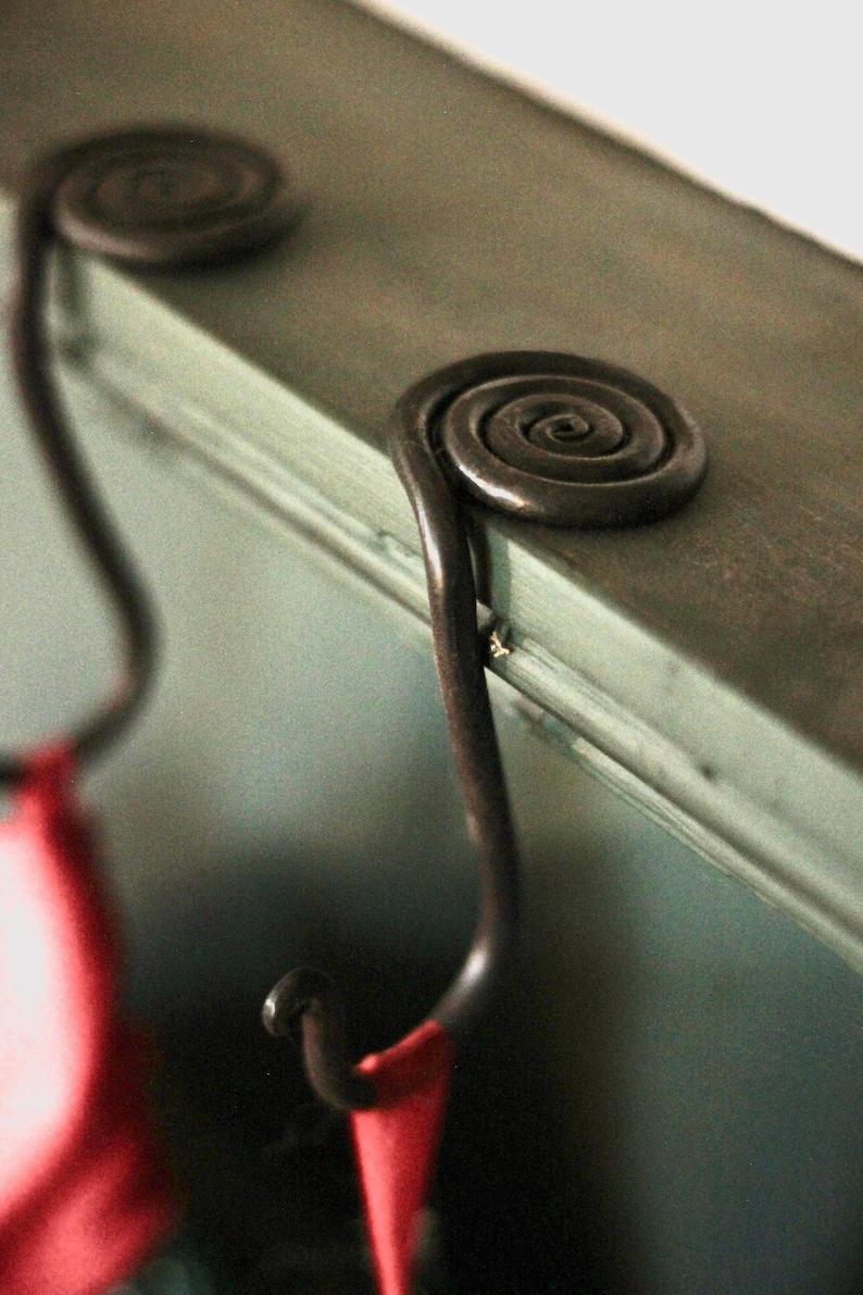 Puede incluir: Un gancho de metal negro con un dise&ntilde;o en espiral en la parte superior. El gancho est&aacute; unido a una superficie verde y una tela roja cuelga del gancho.