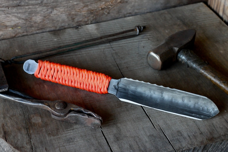 May include: Handmade knife with a textured, dark blade and an orange wrapped handle. The knife is displayed on a wooden surface, alongside blacksmithing tools, including a hammer and tongs. The blade has a serrated edge.