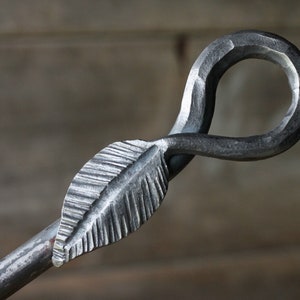 May include: A metal fireplace poker with a leaf-shaped handle and a loop at the end. The poker is made of a dark gray metal and has a rustic finish.