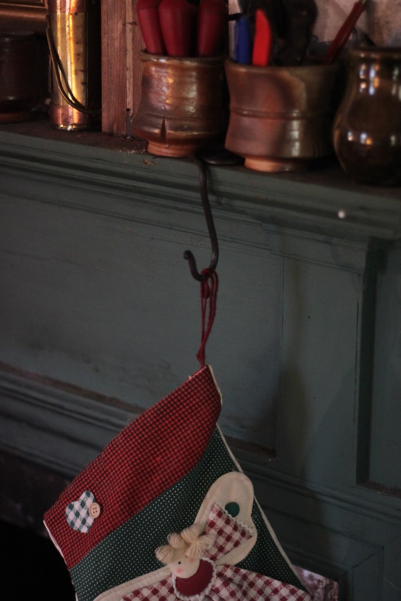Puede incluir: Una media de Navidad roja y verde con un dise&ntilde;o de &aacute;ngel blanco cuelga de un gancho negro en una pared verde oscuro.