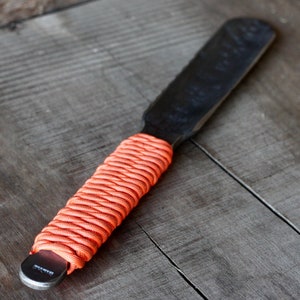 May include: A black metal spatula with a textured blade and an orange paracord-wrapped handle. The spatula is designed for spreading or scraping, and the handle provides a secure grip. The tool is shown on a weathered wooden surface.