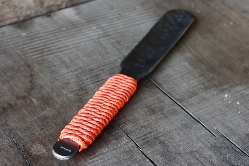 May include: A black metal spatula with a textured blade and an orange paracord-wrapped handle. The spatula is designed for spreading or scraping, and the handle provides a secure grip. The tool is shown on a weathered wooden surface.
