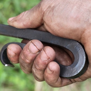 May include: A hand holding a black metal hook with a rounded end. The hook is made of forged steel and has a rustic finish.