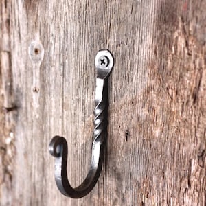 May include: A dark gray, hand-forged metal wall hook featuring a twisted central shaft and a curled end. The hook is secured to a vertical surface with a single black screw through a circular mounting plate at the top of the hardware piece.