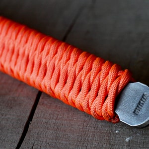 May include: Close-up of a bottle opener with an orange paracord-wrapped handle. The handle is tightly woven, providing a textured grip. The metal opener has a gray finish and the word "FUSTA" is stamped on the end.