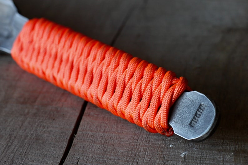 May include: Close-up of a bottle opener with an orange paracord-wrapped handle. The handle is tightly woven, providing a textured grip. The metal opener has a gray finish and the word "FUSTA" is stamped on the end.