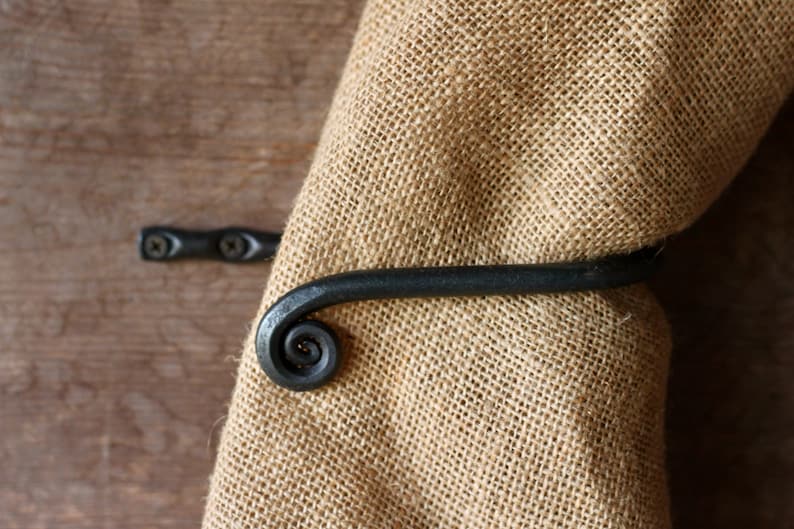 May include: Black metal towel ring with a spiral design. The ring is mounted on a wooden surface and a burlap towel is hanging on it.
