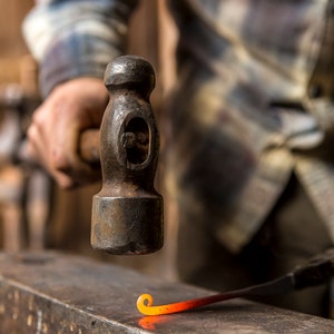 May include: A blacksmith uses a hammer to shape a piece of hot metal on an anvil. The metal is glowing orange and is being bent into a curved shape.