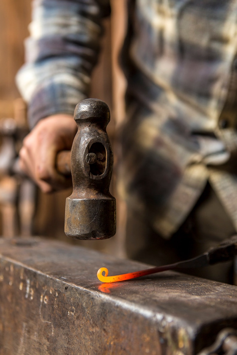 May include: A blacksmith uses a hammer to shape a piece of hot metal on an anvil. The metal is glowing orange and is being bent into a curved shape.