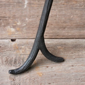 May include: Black metal tripod stand with a forked base. The stand is made of wrought iron and has a rustic, industrial look.