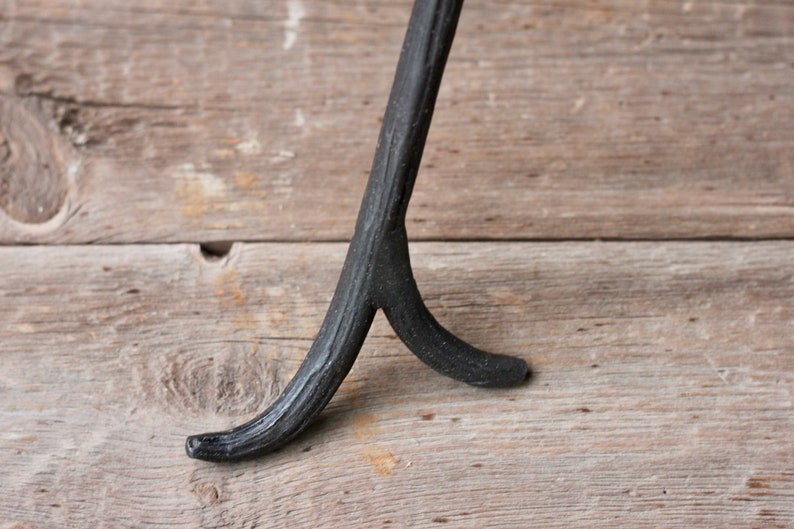 May include: Black metal tripod stand with a forked base. The stand is made of wrought iron and has a rustic, industrial look.