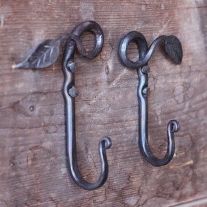 May include: Two black metal hooks with leaf accents mounted on a wooden surface. The hooks are curved and have a decorative swirl at the top.