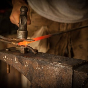 May include: A blacksmith uses a hammer to shape a piece of hot metal on an anvil. The metal glows orange-red as it is worked.