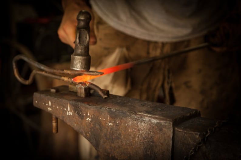 May include: A blacksmith uses a hammer to shape a piece of hot metal on an anvil. The metal glows orange-red as it is worked.