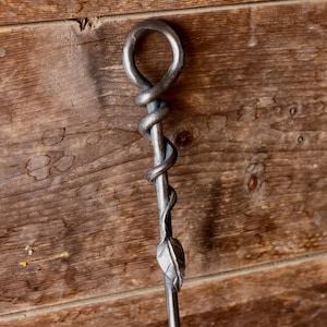 May include: A metal fireplace poker with a twisted, snake-like design and a leaf-shaped tip. The poker is hanging on a wooden wall.