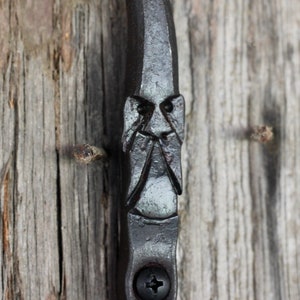 May include: A black metal door handle with a face carved into it. The handle is attached to a wooden door.