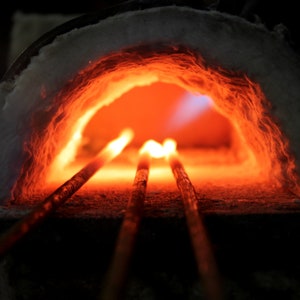 May include: Three metal rods are heated in a forge, glowing orange and red. The forge is lined with white insulation and the rods are partially visible in the opening.