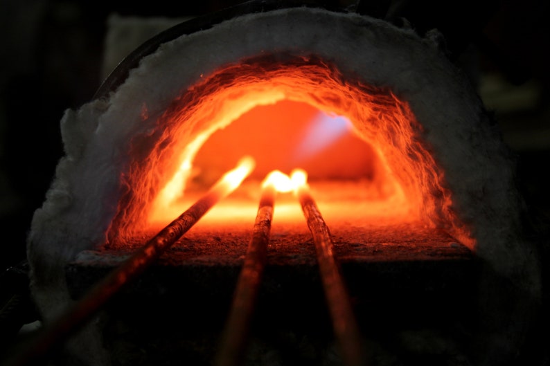 May include: Three metal rods are heated in a forge, glowing orange and red. The forge is lined with white insulation and the rods are partially visible in the opening.