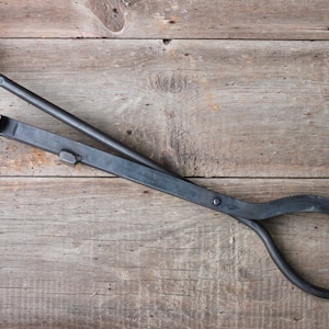 May include: Black metal fireplace tool with a curved handle and a long, straight shaft. The tool is designed to grab and move logs in a fireplace.