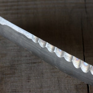 May include: Close-up of a metal tool with a scalloped edge. The tool has a curved shape and a dull, silver finish. The edge has a series of semi-circular cutouts. The background is a wooden surface.