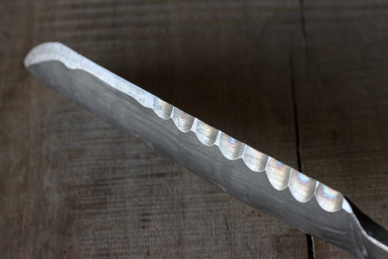 May include: Close-up of a metal tool with a scalloped edge. The tool has a curved shape and a dull, silver finish. The edge has a series of semi-circular cutouts. The background is a wooden surface.