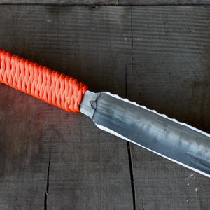 May include: A handmade knife with a textured, silver-colored blade and an orange woven handle. The blade has a serrated edge on one side. The knife rests on a dark wooden surface.