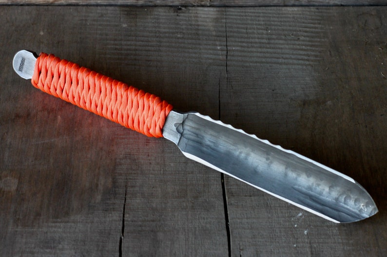 May include: A handmade knife with a textured, silver-colored blade and an orange woven handle. The blade has a serrated edge on one side. The knife rests on a dark wooden surface.