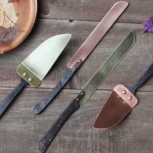 May include: Four handmade cake servers with black handles and different colored metal blades. The blades are copper, brass, and gold.